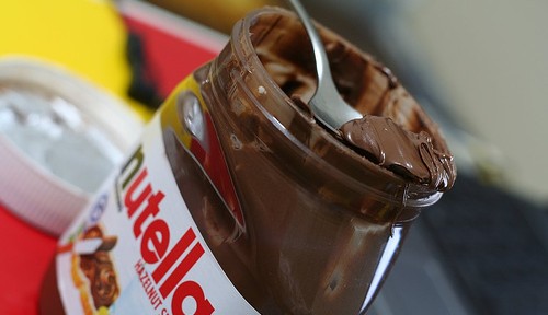 A jar of Nutella with a spoon covered in chocolate hazelnut spread rests inside. The lid is off and placed nearby. The background is slightly blurred, showing a mix of red and yellow colors.