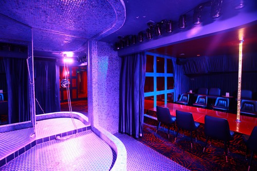 A dimly lit room with a small stage surrounded by mirrors. There's a pole on the stage, and a row of cushioned chairs facing it. The lighting is primarily purple and blue, creating a nightclub-like atmosphere.