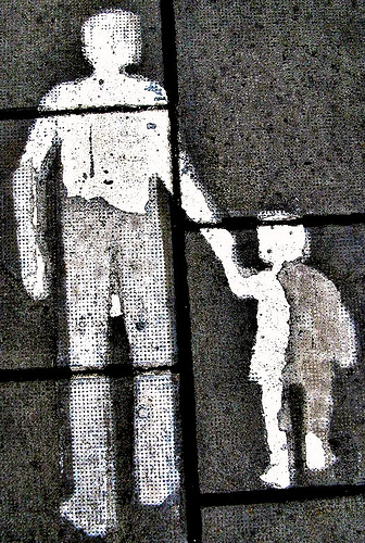 Street art on pavement depicting a white silhouette of an adult holding hands with a child. The figures are painted on gray concrete tiles, suggesting a guiding or protective gesture.