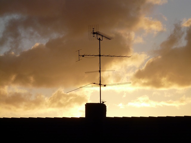 A rooftop TV antenna is silhouetted against a dramatic sunset sky with clouds, creating a striking contrast between the structure and the warm background hues of orange and yellow.
