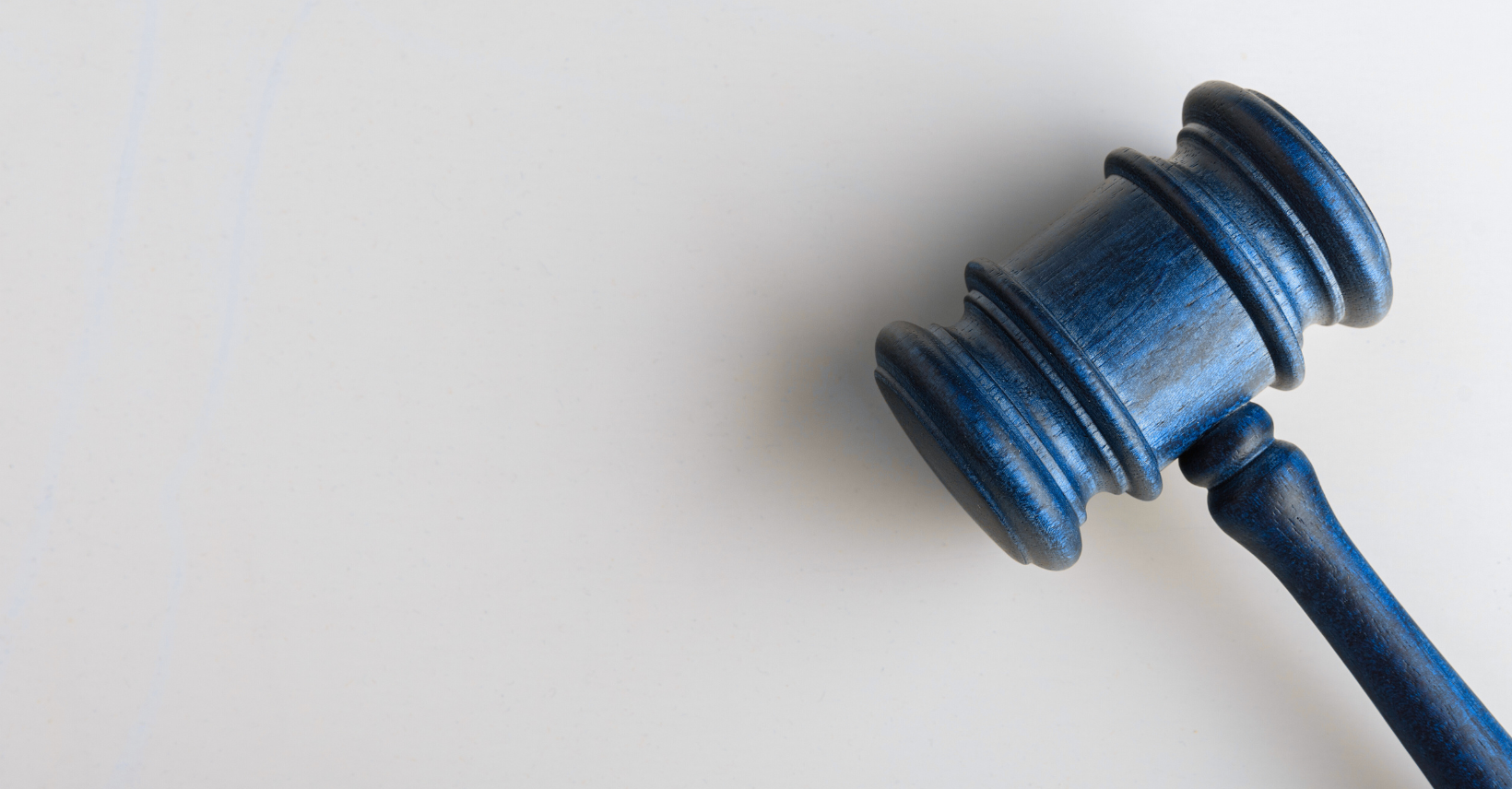 A blue wooden judge’s gavel rests on a light, plain surface, positioned diagonally with the handle pointing downward to the right.