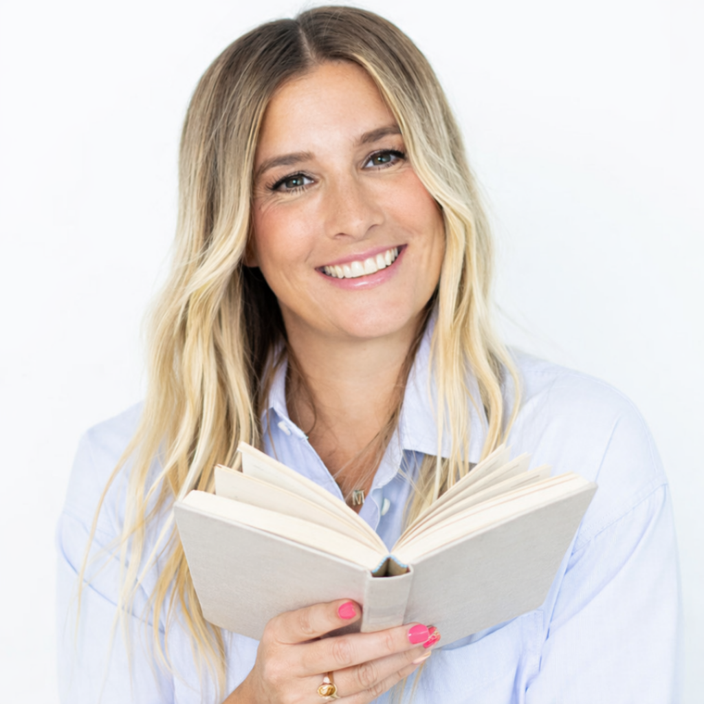 A smiling woman with long blonde hair holds an open book in her hands. She is wearing a light blue shirt and has pink nail polish. The background is plain white.